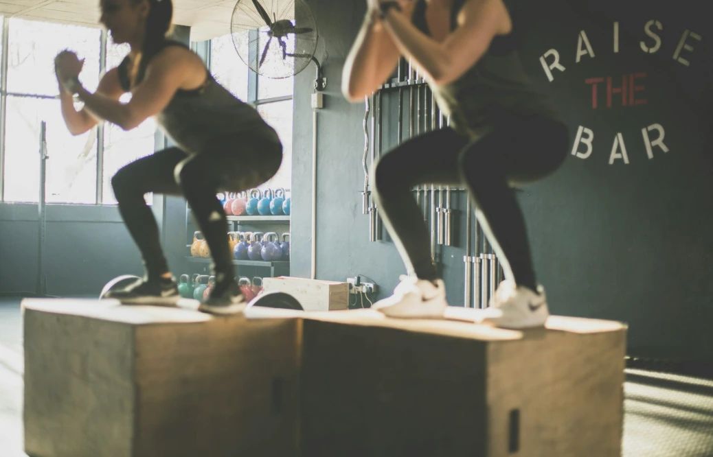 Two people performing box jumps on wooden crates in a gym